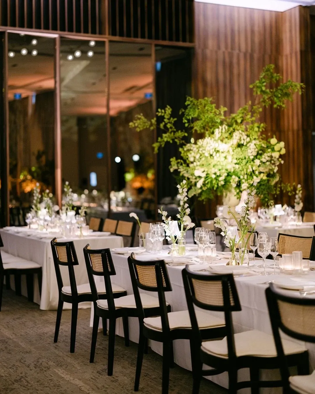 The image shows an elegantly set table at the Westbury Ballroom, featured under the Wedding Venues category on Mariage Elegant, Canada. The ballroom is beautifully decorated with long white tables adorned with delicate floral centerpieces featuring lush greenery and white blooms. Chic black chairs with woven backs are neatly arranged around the tables, and soft ambient lighting creates a romantic atmosphere. The background reveals large mirrors and wooden paneling, adding to the venue’s sophisticated and modern decor, perfect for an elegant wedding celebration.