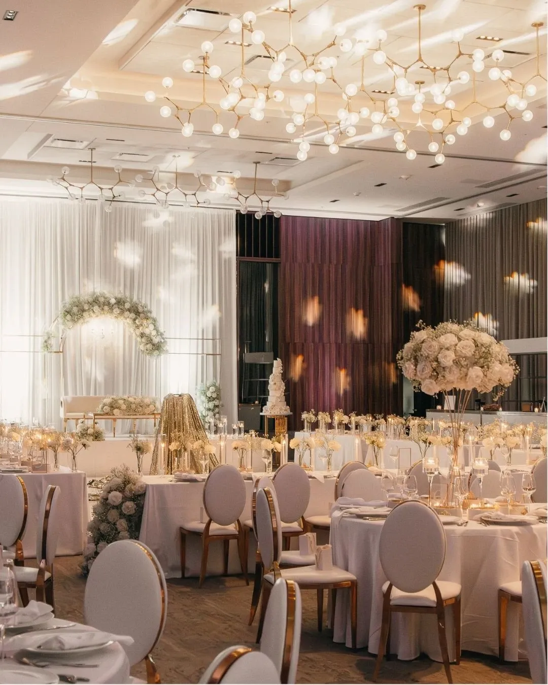 The image showcases the Westbury Ballroom, a wedding venue, elegantly arranged for a wedding reception. The spacious room features round tables adorned with white tablecloths and chic white chairs. Centerpieces of lush white flowers and tall candle arrangements add a touch of sophistication. A beautiful floral arch is present at the front, suggesting a ceremonial or focal point. Soft, ambient lighting creates a romantic atmosphere, enhanced by modern chandeliers overhead. This setting highlights the Westbury Ballroom's capacity to host stylish and memorable weddings.
