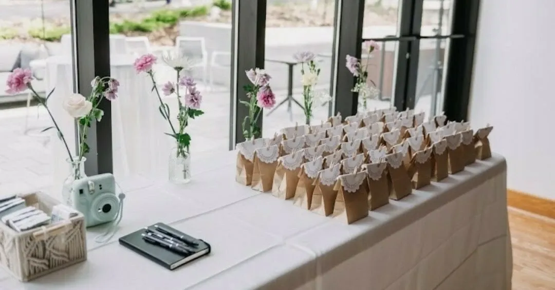 This image features a beautifully arranged wedding planning setup by RLI Events, categorized under Wedding Planners. The scene includes a table adorned with kraft paper gift bags, each topped with delicate lace, likely prepared for wedding guests. A charming collection of pink and white flowers in clear glass vases adds a soft, romantic touch to the table. Additionally, a vintage-style instant camera and a black notebook with pens suggest preparations for capturing special moments and notes. The setup is beside large windows, allowing natural light to illuminate the space, enhancing the elegant and welcoming ambiance suitable for a wedding event.