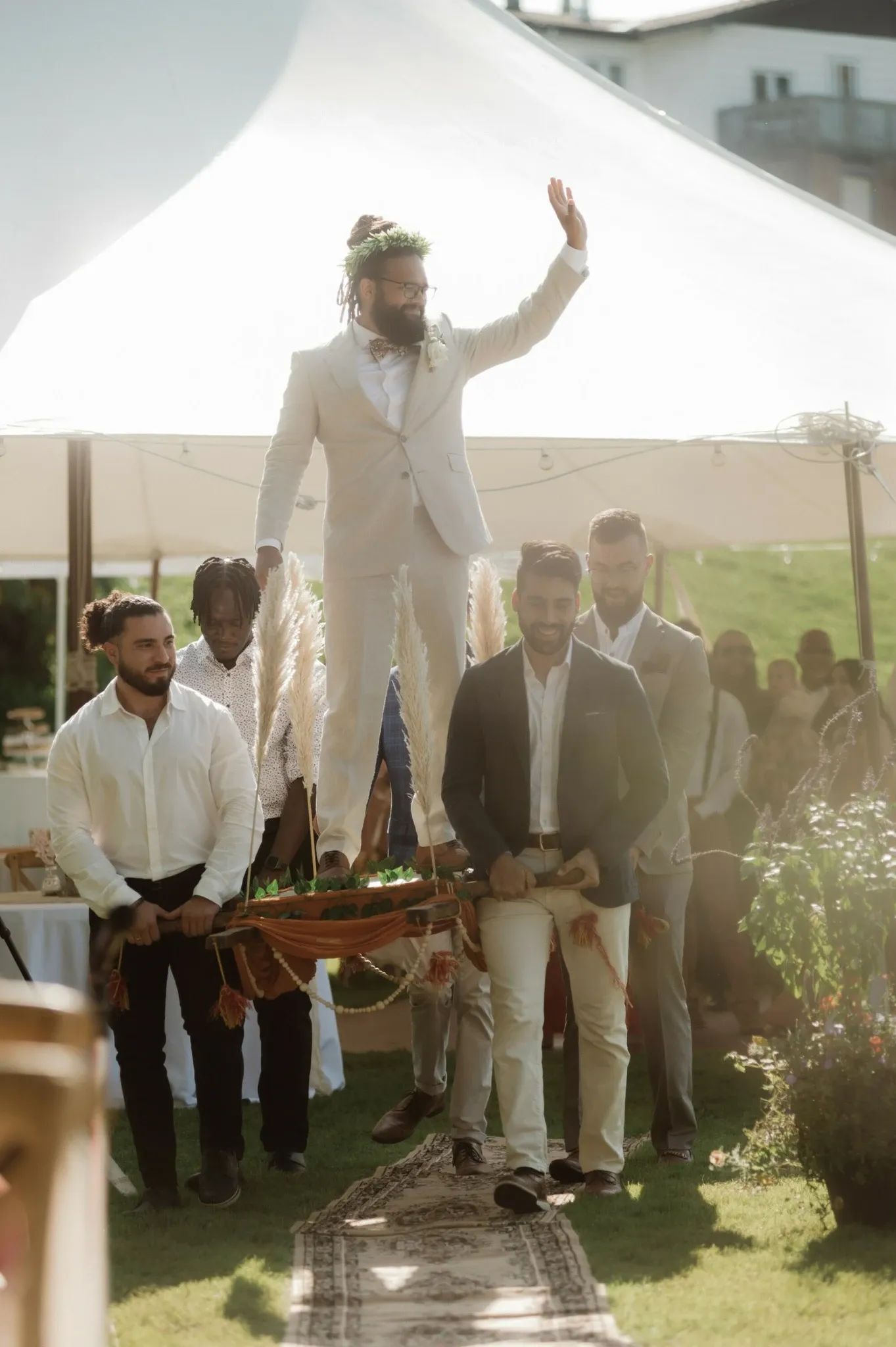 This image features a groom in a light-colored suit with a floral crown, joyfully raising his hand while standing on a traditional wooden palanquin. The palanquin is carried by several groomsmen dressed in white shirts and dark blazers, creating an elegant and festive atmosphere. The backdrop displays a white tent, adding to the outdoor wedding setting. This lively scene exemplifies the unique and personalized wedding planning services offered by RLI Events, specialists in creating unforgettable wedding experiences. The image captures an important moment in a celebration, aligning with the Wedding Planners category on Mariage Elegant, Canada.