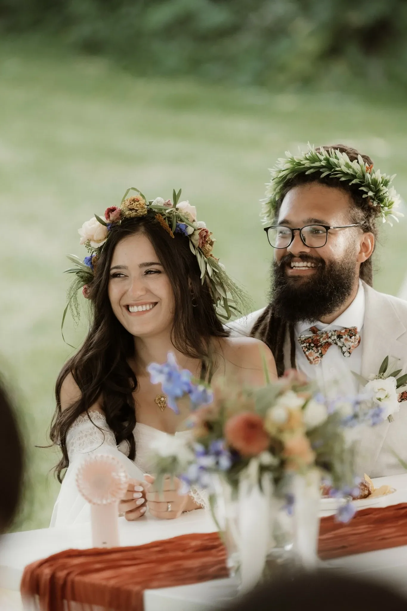 In this image, a joyful couple is seated at a beautifully decorated wedding table outdoors. They are both wearing floral crowns, adding a natural and elegant touch to their attire. The woman is dressed in a white lace wedding gown, while the man is in a light-colored suit with a floral-patterned bow tie that complements their crowns. In front of them, a table holds an assortment of colorful floral arrangements that enhance the romantic atmosphere. This scene captures a moment of happiness, reflecting the couple's joy on their special day. This wedding was organized by RLI Events, showcasing their expertise in creating enchanting and memorable occasions as highlighted in the Wedding Planners category on Mariage Elegant, Canada.