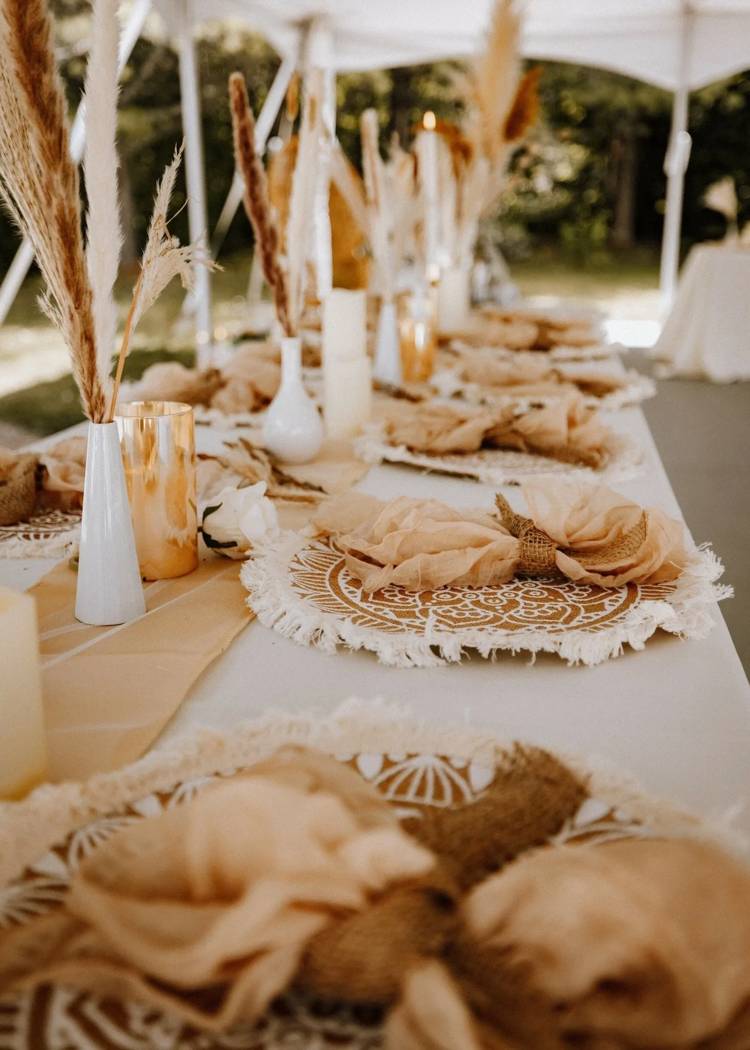 The image showcases an elegantly arranged outdoor wedding reception setup by RLI Events, under the category of Wedding Planners on Mariage Elegant, Canada. A long table is adorned with intricate lace placemats and beautifully folded beige napkins tied with twine. The table features a variety of decor, including tall, slender vases with dried pampas grass, adding a rustic and sophisticated touch. Soft candlelight from strategically placed candles creates a warm and inviting ambiance, perfect for a memorable wedding celebration. The natural outdoor setting enhances the romantic atmosphere, ideal for a stylish and unique wedding experience.
