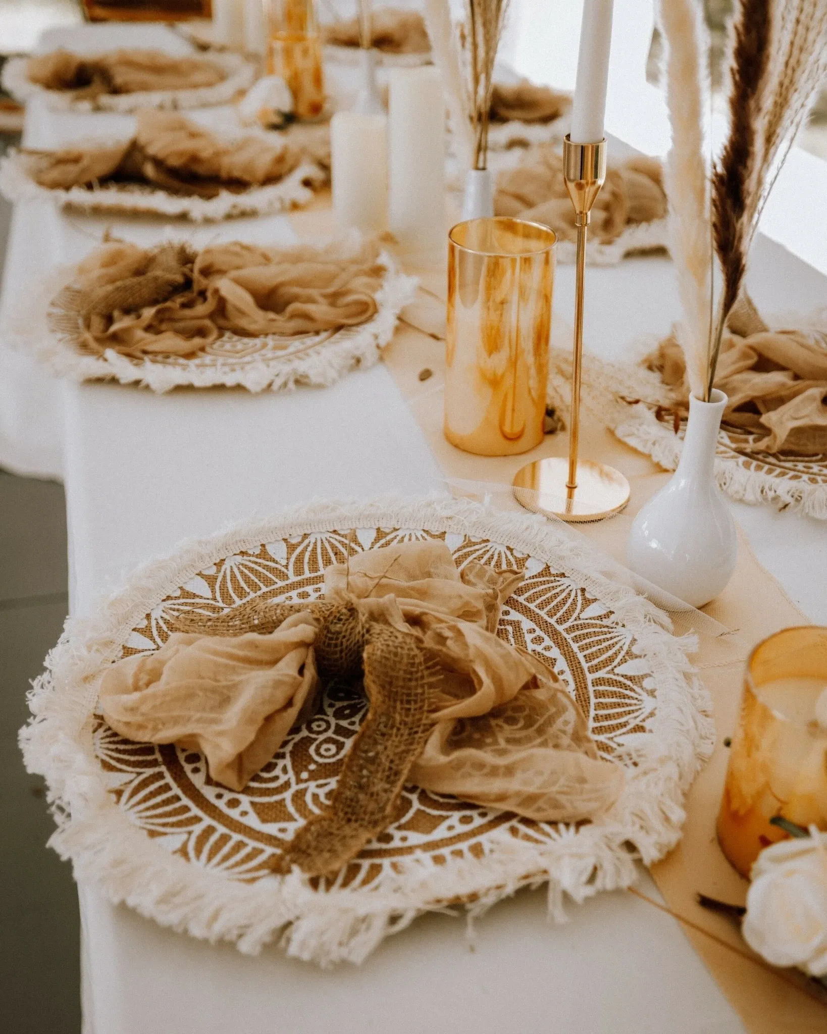 The image showcases a beautifully decorated wedding table set by RLI Events, a wedding planner in the Mariage Elegant vendor listing. The table features intricately designed placemats with a lace-like pattern, complemented by soft, beige napkins artistically tied at the center. Elegant gold and white accents, including tall candles and delicate vases with pampas grass, add a touch of sophistication. The arrangement combines rustic charm with modern elegance, ideal for a stylish wedding reception.