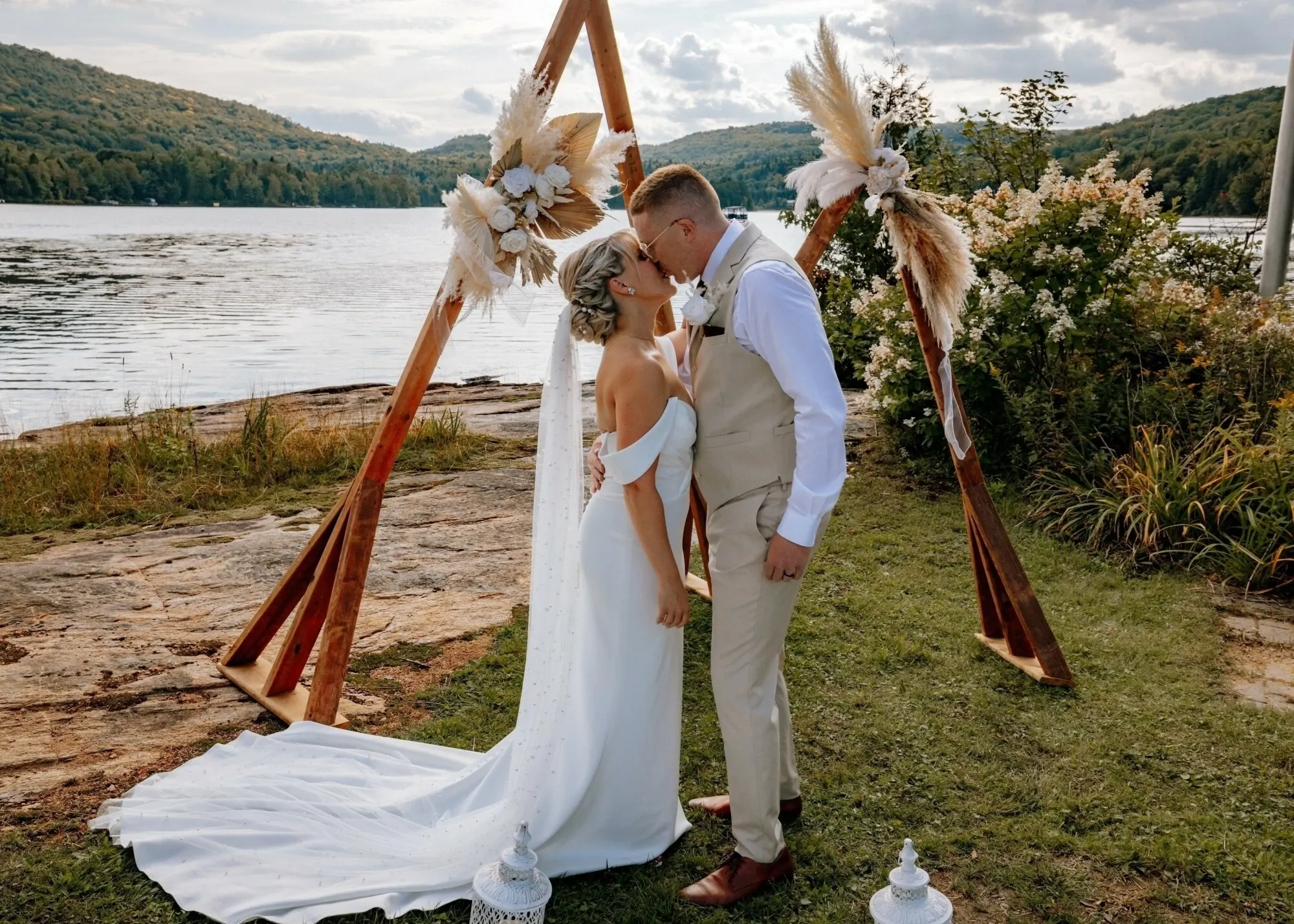 A bride and groom stand in front of a triangular wooden wedding arch decorated with pampas grass and white flowers by the lakeside. The bride is wearing an elegant white off-the-shoulder gown with a long train, while the groom is dressed in a beige vest and trousers with a white shirt. They are sharing a romantic moment in a picturesque outdoor setting, surrounded by lush greenery and hills in the background. This image represents the work of RLI Events, a wedding planner in Canada, showcasing their ability to create stunning wedding environments.