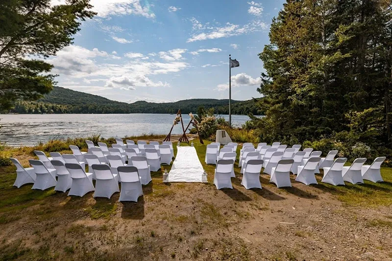 This image showcases a beautifully arranged outdoor wedding ceremony set up by RLI Events, a top vendor in the Wedding Planners category on Mariage Elegant, Canada. The scene features rows of white-covered chairs on either side of a white aisle runner, perfectly aligned and facing a decorative wooden arch. The surrounding natural landscape includes a serene lake and lush greenery under a partly cloudy sky, creating a picturesque and tranquil backdrop ideal for a wedding ceremony.