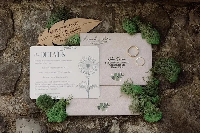 This image features a beautifully arranged flat lay of elegant wedding stationery, perfect for a rustic-themed wedding, provided by RLI Events, a wedding planner in Canada. The scene includes a 'Save Our Date' wooden embellishment and a tastefully designed invitation card, placed on a natural stone surface, surrounded by lush green moss. The invitation card provides essential wedding details including the date and location, adding a personal touch to the presentation. Two gold wedding rings are thoughtfully positioned, symbolizing the upcoming union. This display by RLI Events highlights their expertise in creating stunning and cohesive wedding themes.
