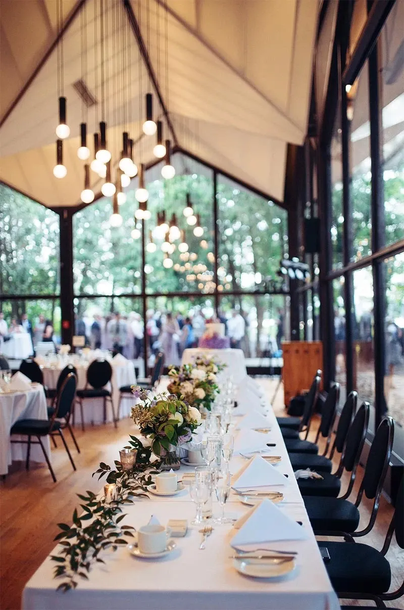 This image showcases a beautifully decorated wedding reception venue, organized by RLI Events, a premium wedding planner in the Mariage Elegant vendor listing in Canada. The spacious room features long, elegantly set tables adorned with white tablecloths and lush greenery centerpieces, creating a classic and sophisticated atmosphere. Overhead, stylish pendant lighting adds a warm ambiance to the space. Large windows offer a view of the scenic outdoors, allowing natural light to flood the venue. The setting reflects the meticulous planning and attention to detail that RLI Events brings to wedding celebrations.