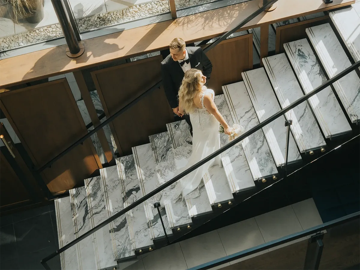 This image showcases a bride and groom elegantly descending a spacious, marble staircase, captured by Muriel Oe Photo, a prominent vendor in the Wedding Photography category for Mariage Elegant, Canada. The bride, in a stunning white gown and holding a bouquet, walks alongside the groom, who is sharply dressed in a black tuxedo. The bright, natural light streaming through the windows highlights the intricate details of the staircase and creates a warm atmosphere, perfect for a memorable wedding photo. The setting suggests a modern, chic venue ideal for a sophisticated wedding celebration, beautifully photographed to capture the couple's special moment.