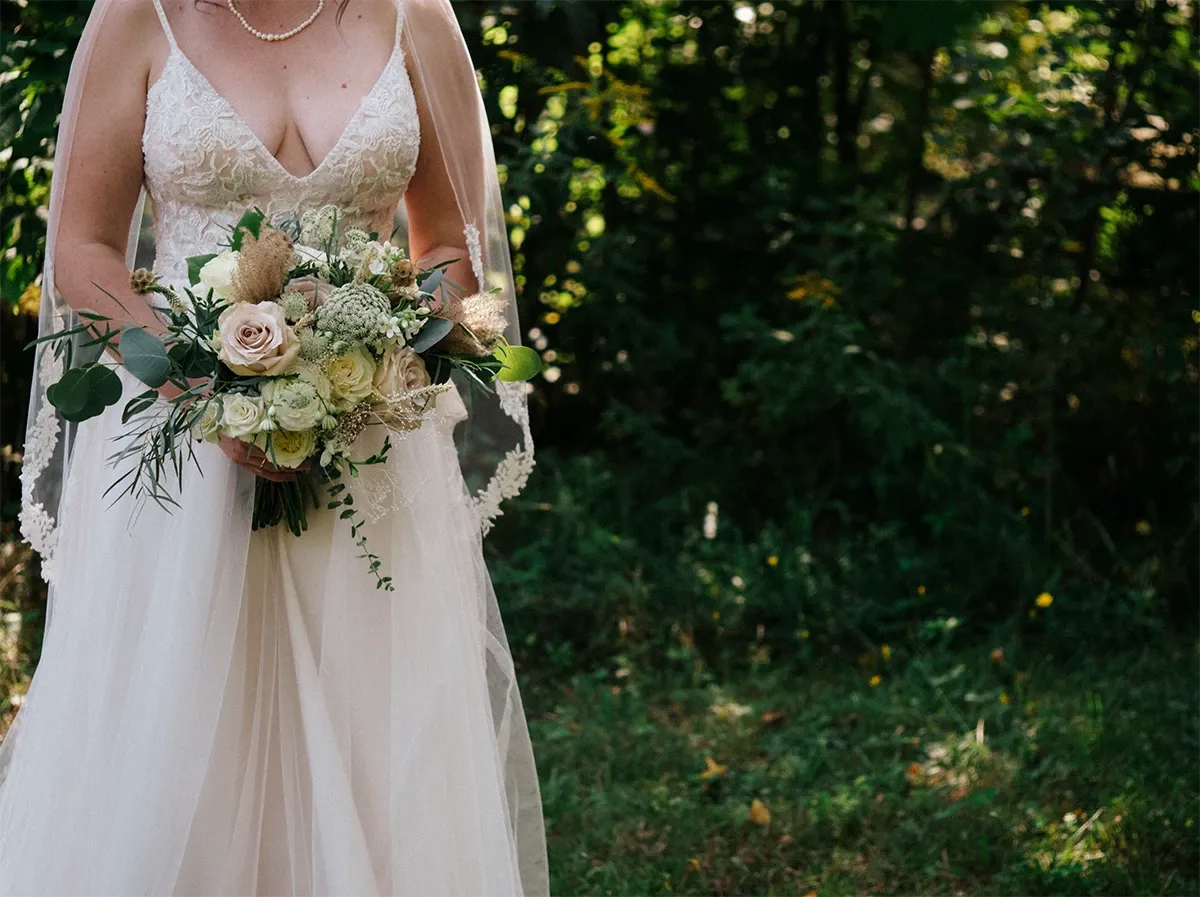 A bride stands in a lush, green outdoor setting, wearing a lace wedding dress with a deep V-neckline, and a delicate necklace. She holds a bouquet featuring white and cream roses, greenery, and pampas grass, adding a natural, elegant touch. The image is captured by Muriel Oe Photo, specializing in wedding photography, and highlights their ability to beautifully showcase intimate wedding moments amidst nature for Mariage Elegant, Canada.