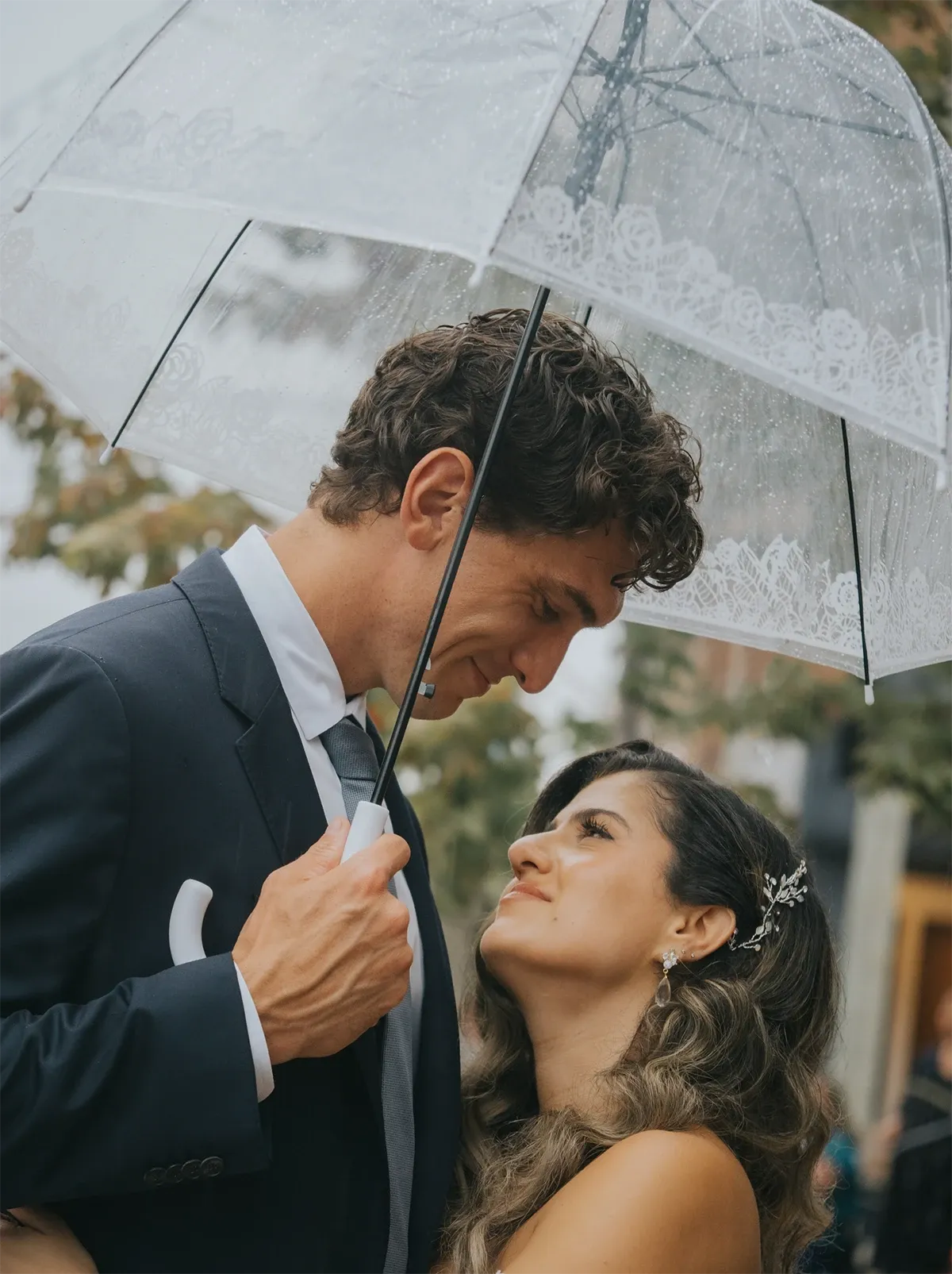 A bride and groom share an intimate moment under a clear, lace-trimmed umbrella, with gentle rain creating a romantic atmosphere. The groom, dressed in a tailored navy suit and grey tie, lovingly gazes down at the bride, who wears an elegant white wedding dress adorned with a decorative hairpiece. The lush green foliage in the background adds a natural touch to the scene. This image, captured by Muriel Oe Photo, showcases the beauty of wedding photography, perfectly aligning with the offerings of Mariage Elegant in Canada.