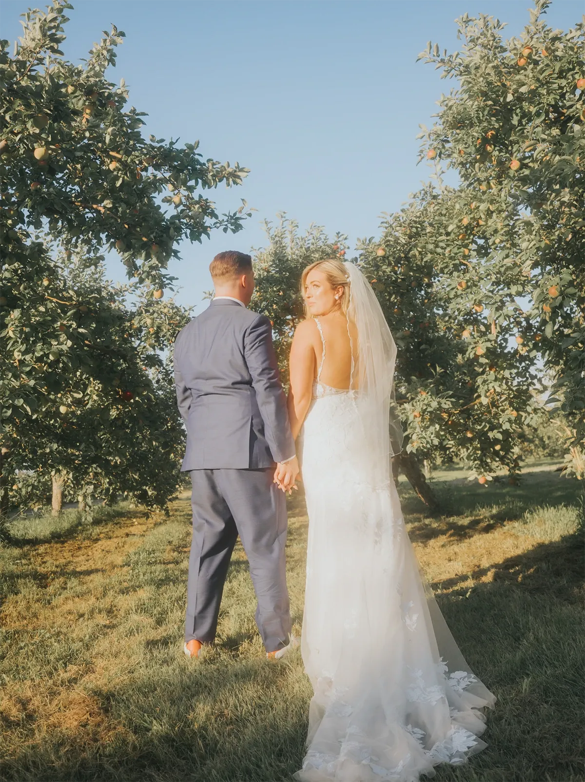 A newlywed couple walks hand in hand through a picturesque orchard under a clear blue sky. The bride is wearing an elegant, lace wedding dress with a flowing veil, while the groom is dressed in a sharp navy suit. Sunlight gently filters through the leaves, creating a romantic atmosphere. Captured by Muriel Oe Photo, this image exemplifies the enchanting moments of a wedding day, showcasing the beautiful natural setting often highlighted in the wedding photography category by Mariage Elegant, Canada.