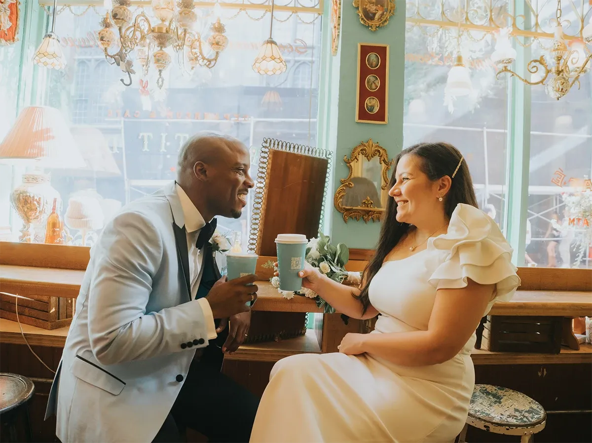 This image by Muriel Oe Photo, a vendor in the Wedding Photography category, captures a joyful couple sharing a moment in a charming café setting. The groom is dressed in a light blue tuxedo with a black bow tie, and the bride is wearing an elegant white dress with ruffled sleeves. Both are smiling warmly at each other while holding coffee cups. The background features a beautifully decorated interior with vintage-style lamps and a large, ornate mirror, adding a romantic and intimate atmosphere, perfect for wedding photography.