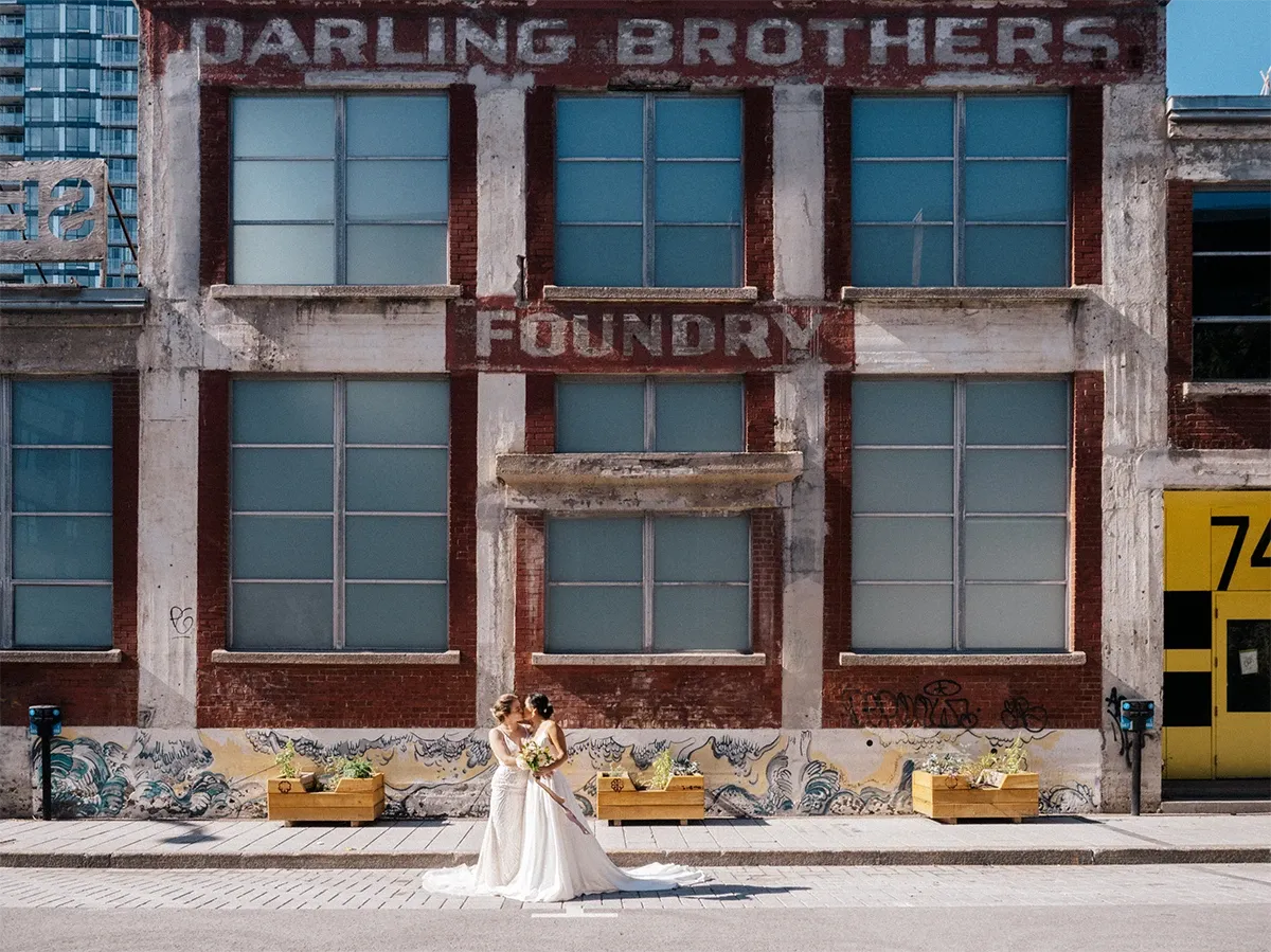 This image from the wedding photography category showcases a beautiful scene captured by Muriel Oe Photo. It features two brides standing lovingly together in front of an industrial-style building with large windows and brick walls, adding an urban twist to their wedding day. The words 'Darling Brothers Foundry' are prominently displayed on the building above them. The brides are elegantly dressed in light-colored gowns and are holding vibrant floral bouquets. The background graffiti art and large wooden planters with greenery introduce a modern, artistic vibe to the photograph, reflecting the unique appeal of urban wedding settings.