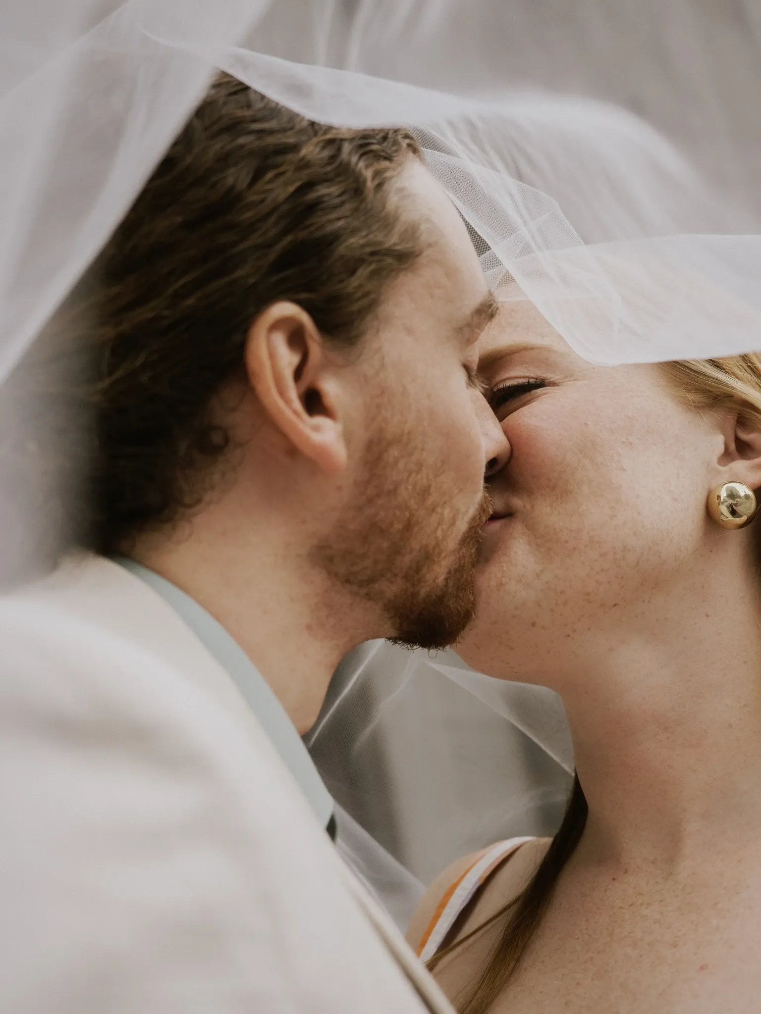 A bride and groom share a tender kiss under a sheer veil, encapsulating a romantic moment on their wedding day. The bride wears elegant pearl earrings and her hair is styled neatly, complementing her graceful appearance. The groom is dressed in a light-colored suit that adds to the serene atmosphere of the photograph. This image, captured by Muriel Oe Photo, showcases the intimate and emotional moments of a wedding, highlighting their expertise in the Wedding Photography category on Mariage Elegant, Canada.