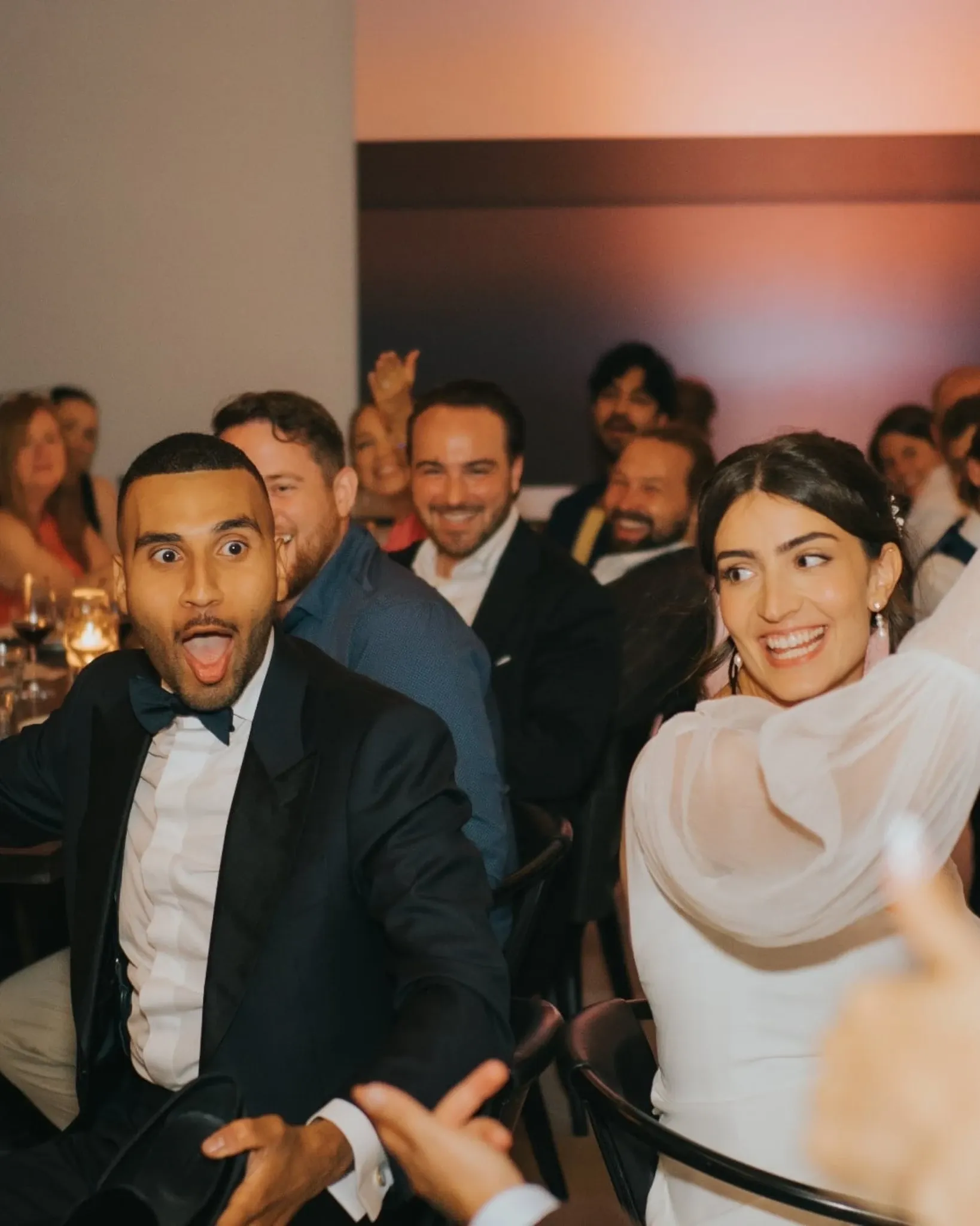At an elegant wedding reception, a joyful couple celebrates amidst a group of guests. The groom, wearing a classic navy tuxedo, displays an expression of surprise and delight, while the bride, in a gracefully designed white dress, beams with happiness. The warmly lit room is filled with well-dressed guests seated at tables adorned with delicate candles. This vibrant moment, captured by Muriel Oe Photo, exemplifies the joyous and candid emotions of a wedding, highlighting the vendor's expertise in wedding photography.