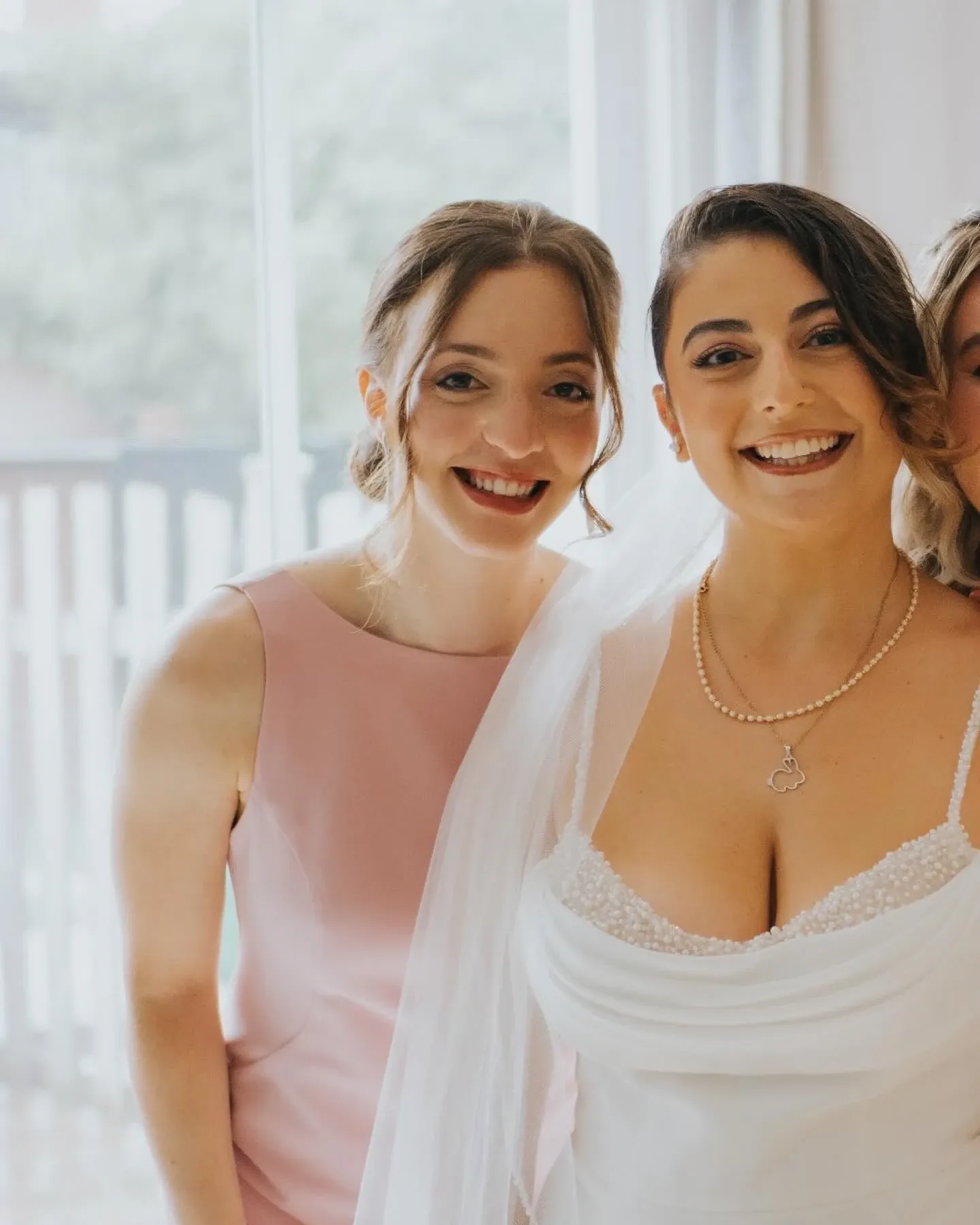 This image features a joyful moment captured by Muriel Oe Photo, a vendor in the Wedding Photography category for Mariage Elegant in Canada. It shows two smiling women, likely part of a wedding celebration. The bride is on the right, wearing an elegant white gown with intricate beadwork and a delicate necklace, complemented by a sheer veil. Beside her, a bridesmaid in a light pink dress shares the moment, standing in front of a softly lit window that reveals a hint of greenery outside. This image beautifully captures the warmth and happiness of a wedding day.