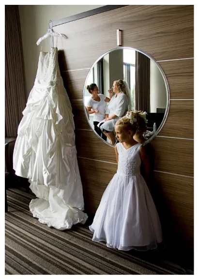 A wedding gown hangs elegantly on a wall-mounted hook beside a large, round mirror in a modern room. In the reflection, a bride is having her makeup applied by a professional artist, showcasing the preparations for her big day. In the foreground, a young flower girl dressed in a white dress with a delicate floral headband gazes towards the window light. This image is taken by Studio Lucie Gamache, highlighting their expertise in capturing intimate and preparatory moments in wedding photography.