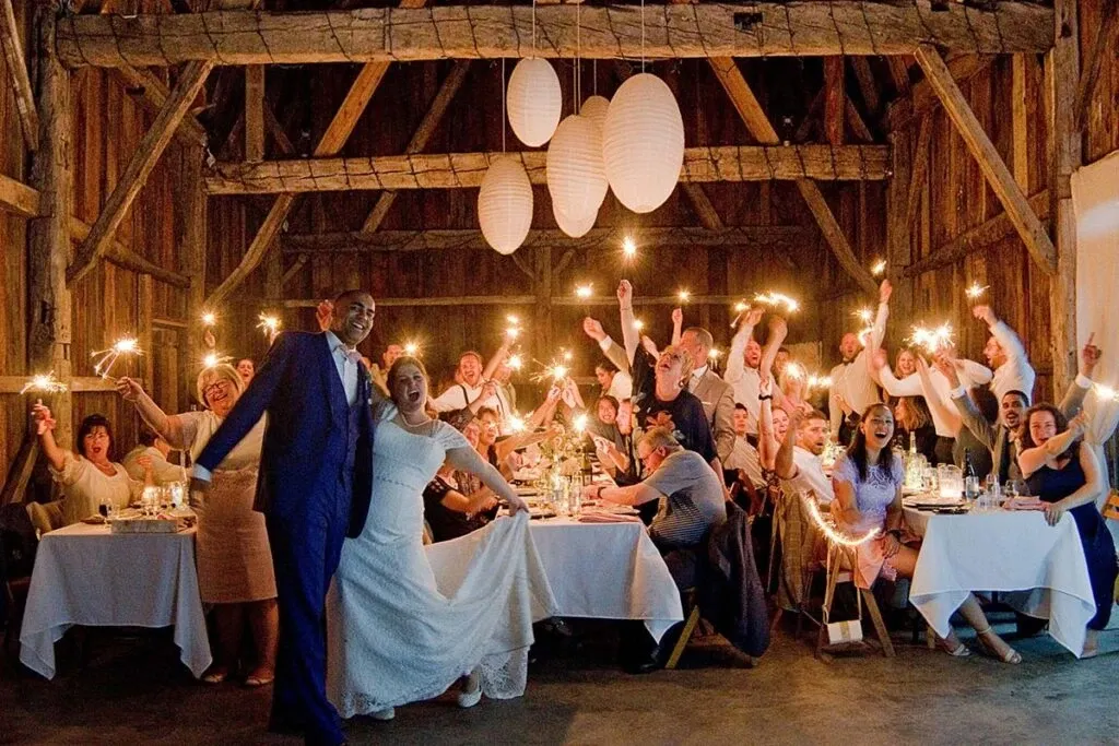 In this image by Studio Lucie Gamache, a vendor specializing in wedding photography, a joyful wedding celebration is captured. The photograph features a newlywed couple dancing at the forefront, with the bride in a white gown and the groom in a blue suit. Behind them, a group of wedding guests sits at decorated tables in a rustic barn setting, holding sparklers aloft. White paper lanterns hang from the ceiling, adding to the festive atmosphere. This lively scene encapsulates the joyous spirit of weddings, making it a perfect illustration for Mariage Elegant in Canada.