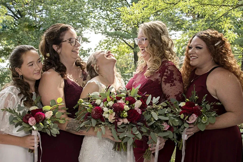 The image shows a joyful bridal party captured by Studio Lucie Gamache, a vendor in the Wedding Photography category for Mariage Elegant, Canada. Five women stand closely together, wearing coordinated burgundy and white dresses, and are holding elegantly arranged bouquets featuring red and pink flowers with green foliage. They are outdoors with lush green trees in the background, all sharing a moment of laughter and happiness, highlighting the positive emotions of a wedding day.