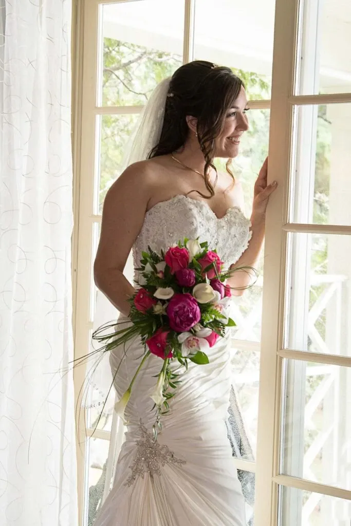 A bride is standing beside a large window with sheer curtains, looking outside with a gentle smile. She is wearing a strapless white wedding gown adorned with intricate lace detailing and a jeweled embellishment at the waist. Her hair is styled elegantly with a veil cascading down her back. In her hands, she holds a vibrant bouquet featuring pink and white flowers with lush greenery, adding a pop of color to the scene. The warm natural light from the window highlights her joyful expression and the exquisite details of her dress. This image showcases the work of Studio Lucie Gamache in the Wedding Photography category for Mariage Elegant, Canada.