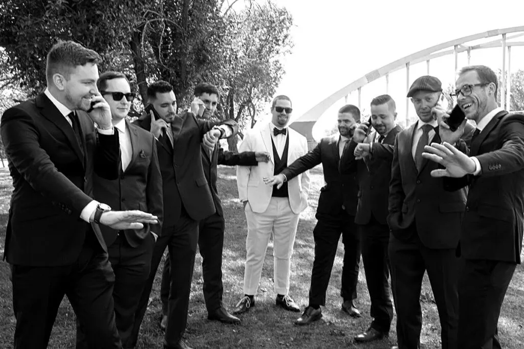 A group of nine men, dressed in formal suits, are standing outdoors on a grassy area under the shade of trees. The men are playfully extending their arms forward, smiling and laughing as they pose for the photo. The groom, wearing a distinctive white suit and bow tie, stands in the center, surrounded by his groomsmen in darker suits. In the background, part of a white arched bridge is visible. This candid moment captures the joyous spirit of a wedding day, photographed by Studio Lucie Gamache, specialists in wedding photography.