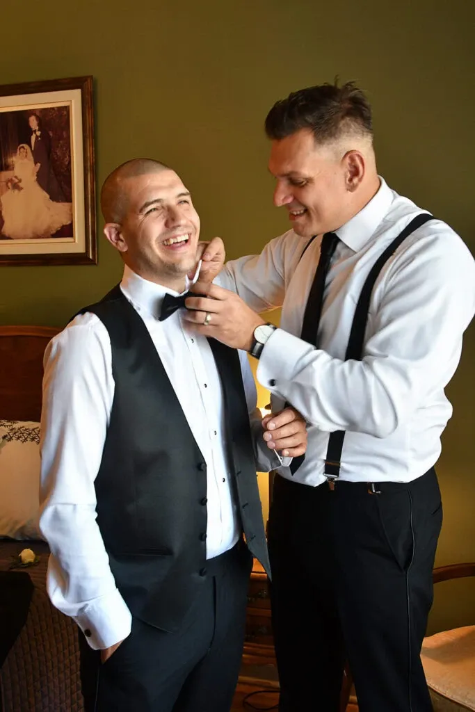In this image from Studio Lucie Gamache, a vendor in the Wedding Photography category, two groomsmen are preparing for a wedding. One man, smiling broadly, is dressed in a crisp white shirt, a black vest, and a bow tie, while the other is helping to adjust his tie. Both men exude a joyful and celebratory atmosphere. They are in a warmly decorated room with a green wall, featuring a framed vintage wedding photograph in the background, capturing the essence of elegance and tradition that is synonymous with weddings in the Mariage Elegant context in Canada.