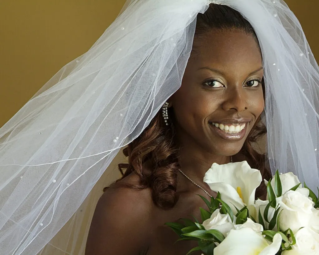 This image showcases a smiling bride on her wedding day, captured by Studio Lucie Gamache, a vendor in the Wedding Photography category on Mariage Elegant, Canada. The bride is wearing a white veil with delicate, pearl-like decorations and holding a bouquet of white calla lilies. Her earrings sparkle as she poses against a soft, neutral background, highlighting her joyful expression on this special occasion.