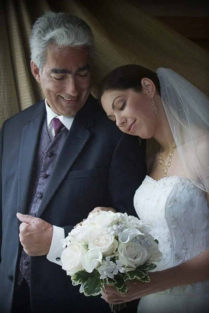 This image features a bride and an older gentleman, likely her father, sharing a tender moment indoors. The bride is dressed in an elegant white wedding gown adorned with delicate embroidery and lace details, paired with a simple yet graceful veil. She is holding a bouquet of fresh white roses interspersed with small white flowers and greenery. The gentleman is wearing a dark suit with a purple vest and a coordinating tie, looking at the bride with a warm, affectionate smile. The photograph, captured by Studio Lucie Gamache, highlights the emotional connection between the bride and her companion, exemplifying the beauty and sentiment of wedding photography for Mariage Elegant in Canada.