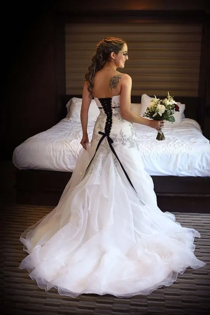 A bride stands gracefully in a well-lit room, captured by Studio Lucie Gamache, a vendor specializing in Wedding Photography for Mariage Elegant, Canada. She is wearing a stunning white gown with intricate black lace detailing on the back, complemented by an elegant flowing train. Her hair is styled in loose curls cascading down her back, and she holds a bouquet of white and deep red flowers, adding a pop of color. The setting includes a neatly made bed in the background, suggesting a moment of calm before the wedding ceremony.