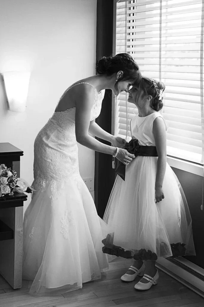 In an elegant black and white photo captured by Studio Lucie Gamache, a wedding photographer listed on Mariage Elegant, Canada, a bride in a stunning lace wedding gown gently touches foreheads with a young flower girl. The flower girl, dressed in a charming white dress with a dark sash, looks up at the bride with a sweet, shared moment of affection. Natural light filters through the window blinds, softly illuminating the intimate scene, which conveys the emotional bond and joyful anticipation of the wedding day.