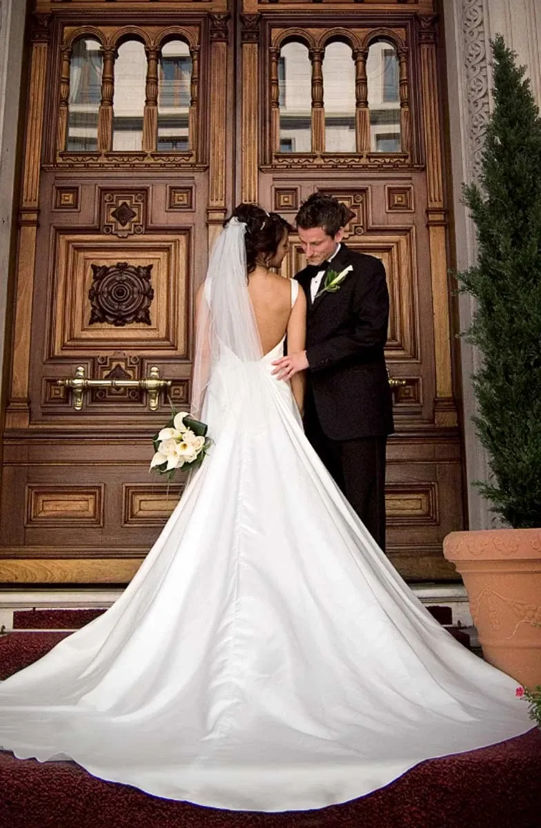 A bride and groom are standing intimately in front of ornately carved wooden doors, captured by Studio Lucie Gamache, a vendor in the Wedding Photography category on Mariage Elegant, Canada. The bride is wearing a flowing white gown with a long train and a veil, holding a bouquet of white flowers. The groom is dressed in a classic black tuxedo, gently resting his hand on the bride's waist. A potted evergreen plant is placed nearby, enhancing the romantic setting.
