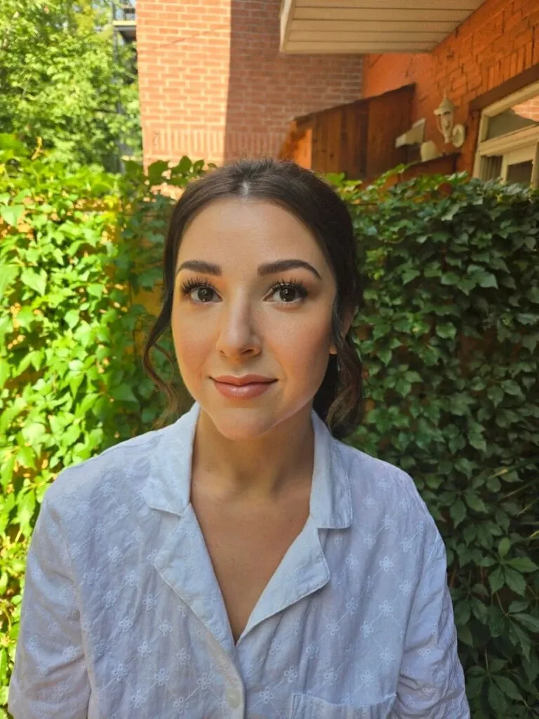 A woman stands outside, surrounded by lush green ivy and brick walls, radiating a fresh bridal look. Her makeup is expertly done with natural tones, emphasizing her eyes with long lashes and subtle eyeliner. Her hair is elegantly styled, complementing the overall sophisticated appearance. She wears a white, delicate-patterned blouse, fitting well with the bridal theme. The setting and style suggest it's part of a professional portfolio for Mariage MTL, a wedding beauty vendor listed on Mariage Elegant, Canada.