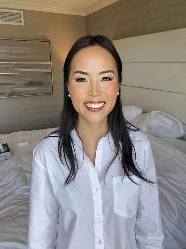 A woman with long, dark hair and expertly applied makeup is smiling warmly at the camera. She is dressed in a crisp, white button-up shirt and stands in a stylish, minimalistic bedroom with neatly arranged bedsheets. This image showcases the bridal beauty services offered by Mariage MTL, under the Wedding Beauty category. The setting provides a glimpse into modern elegance, reflecting the vendor's expertise in enhancing natural beauty for weddings.