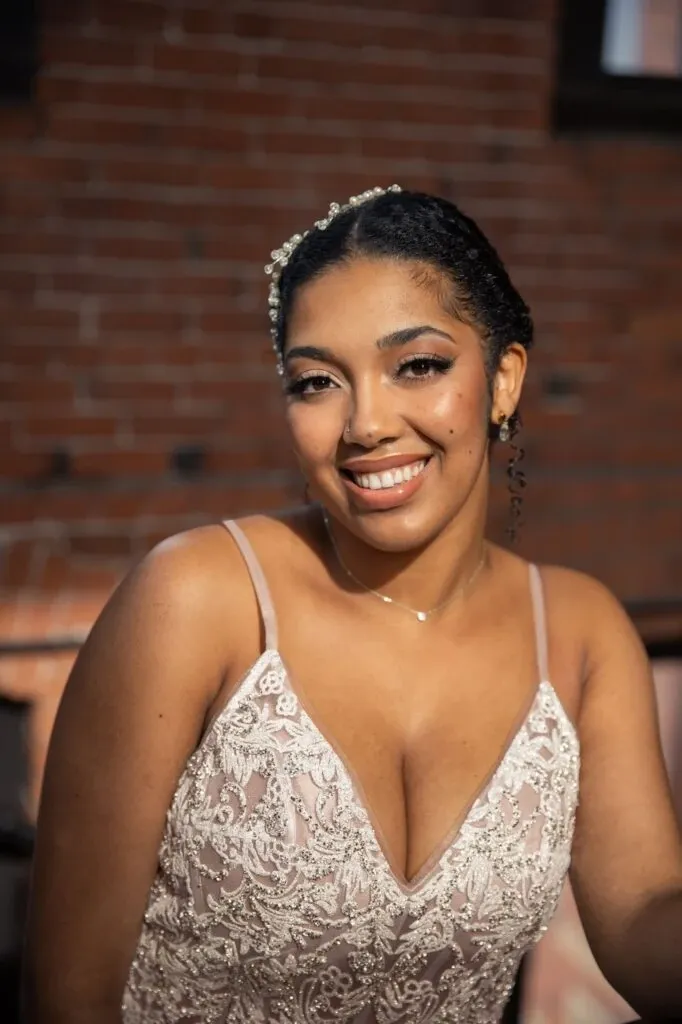 The image shows a smiling bride with elegant makeup and hairstyle, showcasing the wedding beauty services provided by Mariage MTL. Her hair is styled with delicate pearls, complementing her intricate lace bridal gown. The setting features a warm, rustic brick backdrop, adding to the romantic atmosphere. As a representative of the Wedding Beauty category on Mariage Elegant, Canada, this vendor emphasizes elegance and sophistication in bridal beauty.