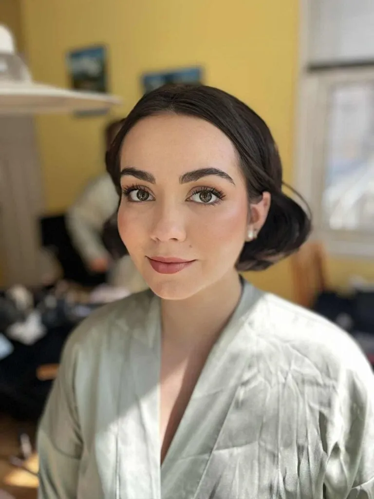 A bride-to-be is getting ready, showcasing an elegant hairstyle and makeup by Mariage MTL, renowned for their expertise in the wedding beauty category. She is wearing a light green satin robe, and her hair is styled in a sleek, classic updo, complemented by soft, natural makeup highlighting her features. The room has a warm, inviting ambiance with a yellow wall in the background and framed pictures subtly visible, enhancing the setting as a preparation space for the wedding ceremony.