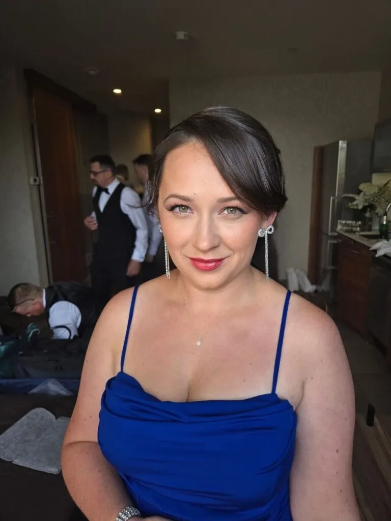 A woman is elegantly styled, showcasing bridal beauty by Mariage MTL in the Wedding Beauty category. She wears a stunning, deep blue spaghetti strap dress, complemented by long, delicate earrings, and subtle makeup that enhances her natural features. Her hair is neatly pinned back, highlighting a classic look perfect for weddings. In the softly lit background, others appear to be preparing, adding to the busy yet joyful atmosphere typical of wedding day preparations. The setting suggests a serene, intimate space, ideal for the bridal party's preparation.