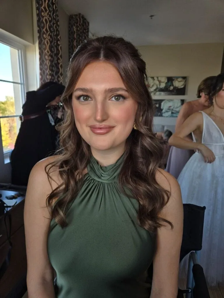 A bride is elegantly seated indoors, showcasing a sophisticated and natural makeup look by Mariage MTL in the Wedding Beauty category. Her long, wavy brown hair is styled half-up, framing her face beautifully. She wears a sleeveless, high-neck green dress, which complements her soft makeup, emphasizing subtle eyeshadow and a nude lip color. In the background, a woman in a white gown is adjusting her attire, adding to the wedding preparation ambiance. A window to the left lets in natural light, highlighting the serene and joyful atmosphere.
