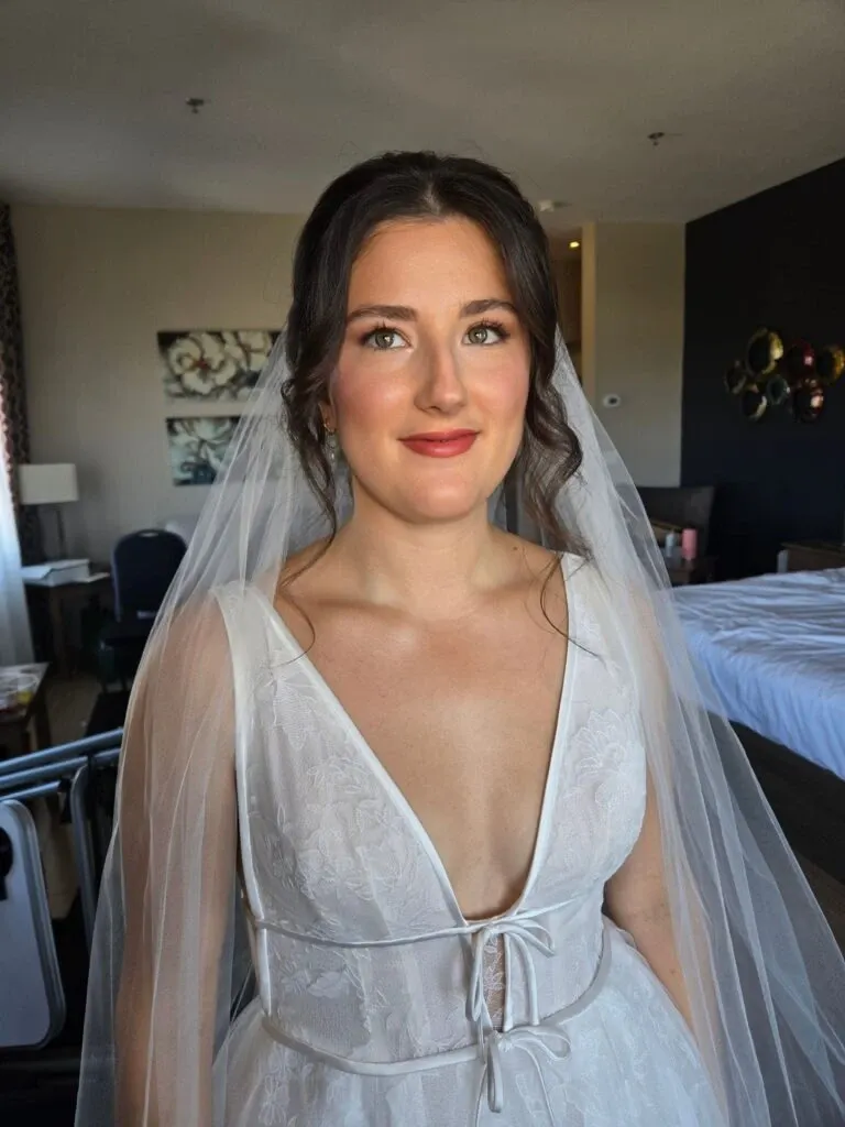 A bride stands in a well-lit room, beautifully showcasing the makeup artistry by Mariage MTL, a vendor in the Wedding Beauty category. She wears a white wedding dress with intricate lace details and a deep V neckline, complemented by a sheer veil cascading down her back. Her makeup is elegantly done with subtle eyeshadow and soft lipstick, enhancing her natural features. The background reveals a tastefully decorated room with modern wall art and neatly arranged furniture, setting a serene and sophisticated atmosphere typical of wedding preparations. This image exemplifies the expertise provided by Mariage MTL within the Mariage Elegant platform in Canada.