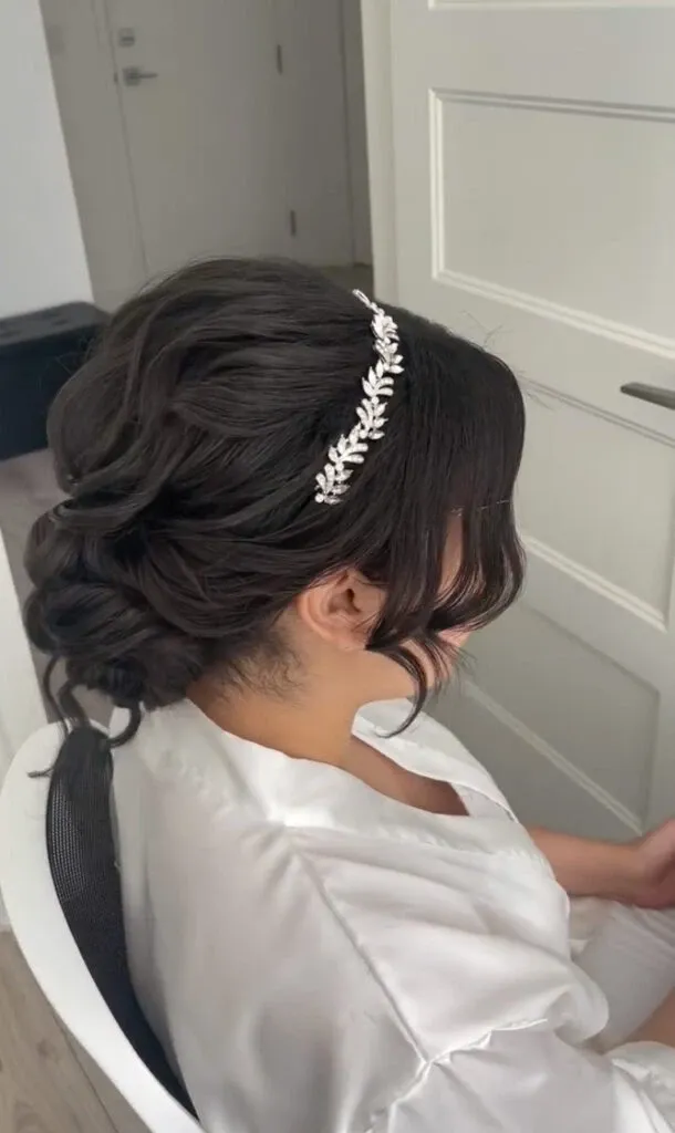 A bride is seated in a white chair, showcasing an elegant updo hairstyle, expertly arranged with soft curls and adorned with a delicate, leaf-patterned headpiece. She is wearing a white satin robe, suggesting preparations on her wedding day. The setting appears to be a bright room with white walls and a partially visible door in the background. The image represents the wedding beauty services offered by Mariage MTL, highlighting their expertise in bridal hairstyling within the wedding category on Mariage Elegant, Canada.