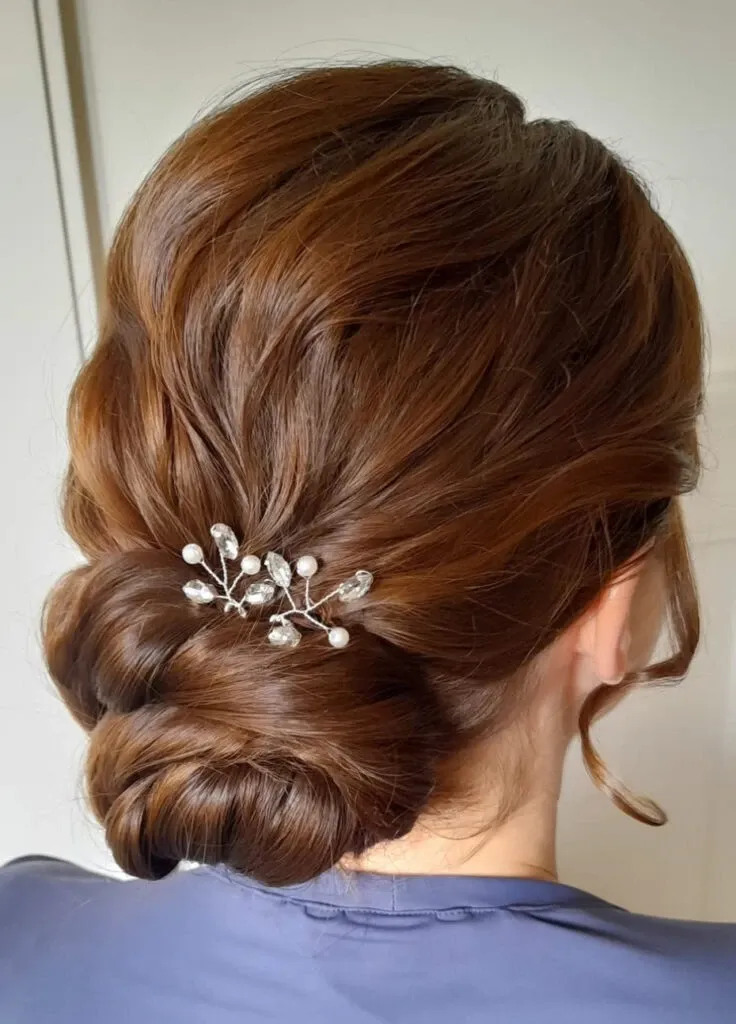 In this image, a bride's elegant hairstyle is showcased, highlighting Mariage MTL's expertise in wedding beauty. The bride's hair is styled in a sophisticated low chignon with soft, smooth waves. The hairstyle is accented with a delicate hairpin featuring small, pearl-like embellishments, adding a touch of classic elegance. This look is perfect for a wedding celebration, emphasizing timeless beauty and attention to detail, which are hallmarks of Mariage MTL's bridal services in Canada.