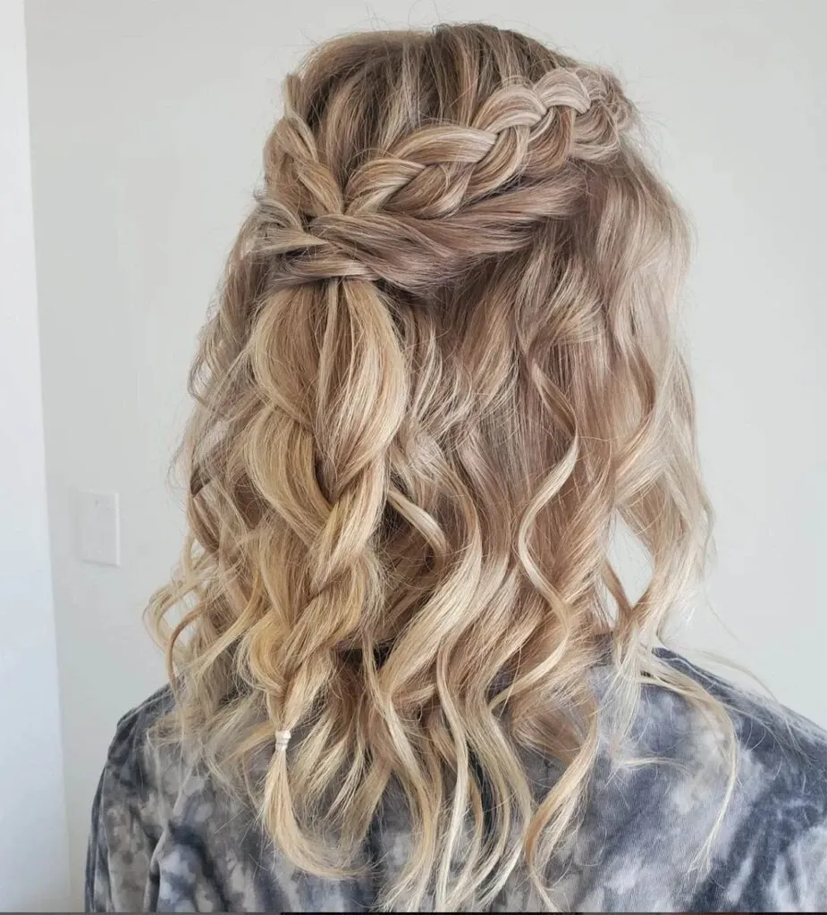 A beautifully styled half-up, half-down hairstyle is showcased, featuring intricate braiding cascading into loose, soft curls. The image highlights wedding beauty expertise by Mariage MTL, a vendor in the Wedding Beauty category. The braided section adds elegance and sophistication, ideal for a bride seeking a romantic, fairytale look. The hair's natural, blond tones create a warm and inviting appearance, perfect for a memorable wedding day in Canada. The backdrop is simple, ensuring the focus remains on the exquisite hair design crafted for a special occasion.