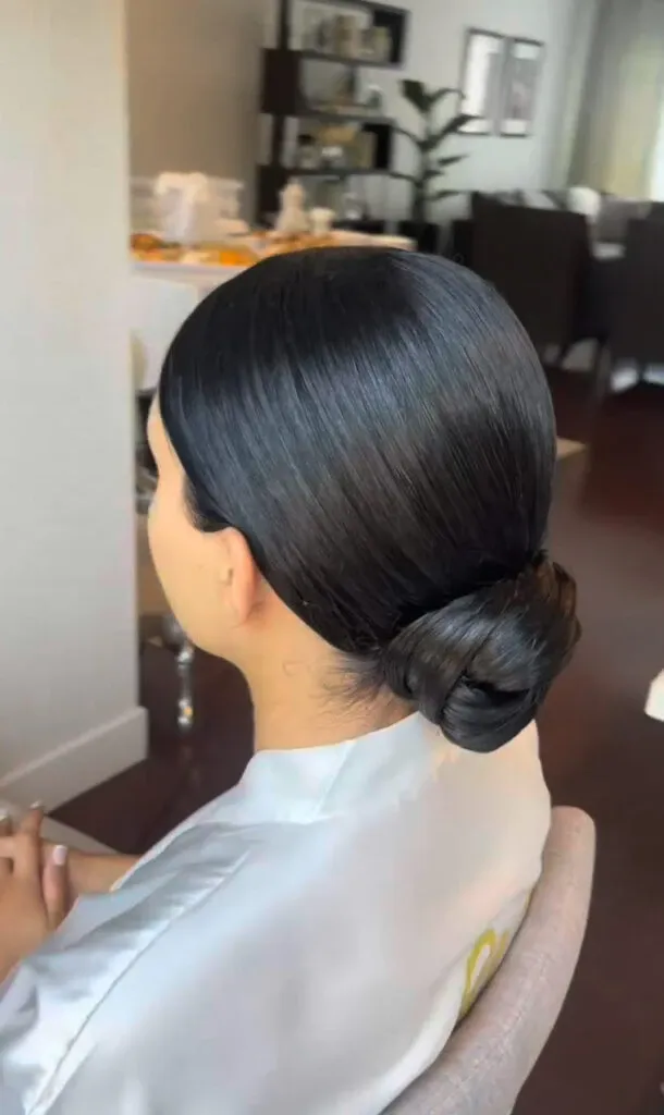 In the image, a woman with sleek, dark hair is styled into an elegant low bun, ideal for a wedding setting. She is wearing a light-colored robe, suggesting she is preparing for a special occasion. The background shows a tastefully decorated room with a table set up, contributing to the sophisticated ambiance. This image showcases the expertise of Mariage MTL in the Wedding Beauty category, highlighting their ability to create refined and polished bridal hairstyles.