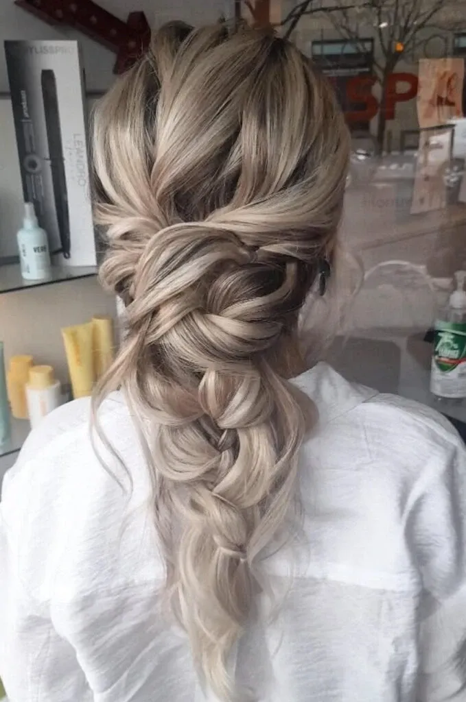 A bride showcasing an elegant hairstyle created by Mariage MTL, a vendor in the Wedding Beauty category. Her long, blonde hair is styled in an intricate braided updo that cascades down her back. The setting appears to be a salon with a professional atmosphere, with shelves in the background displaying various hair care products. This hairstyle exemplifies a sophisticated and timeless look, perfect for a wedding day, highlighting the expertise of the stylist in creating beautiful bridal looks.