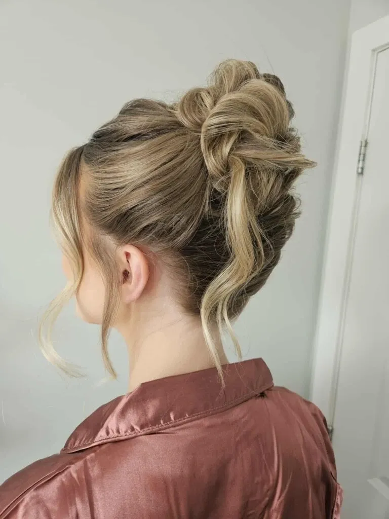 This image showcases an elegant bridal hairstyle created by Mariage MTL, a vendor in the Wedding Beauty category. The focus is on a sophisticated updo featuring intricate twists and delicate waves of blonde hair, beautifully styled to highlight texture and volume. The bride’s hair is adorned with subtle, loose curls gently framing her face, adding a touch of romance to the overall look. She is wearing a silky, blush-colored robe, suggesting a preparation setting, possibly before the wedding ceremony. This style exemplifies the skill and artistry of Mariage MTL, making it a perfect choice for brides seeking refined elegance on their special day.