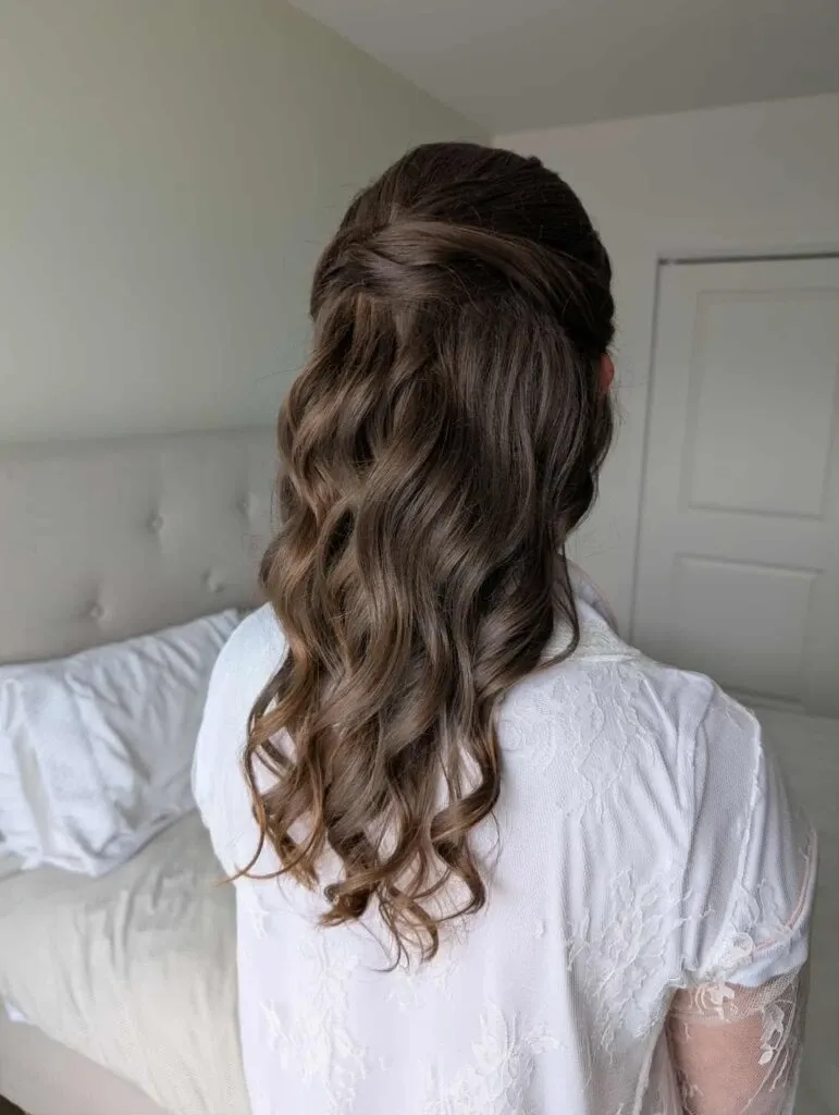 A bride is wearing a lace robe while showcasing an elegant bridal hairstyle, featuring loosely cascading curls and a softly twisted half-up style. The setting is a softly lit bedroom, hinting at bridal preparations. This image is associated with Mariage MTL, a vendor specializing in the Wedding Beauty category for Mariage Elegant in Canada.