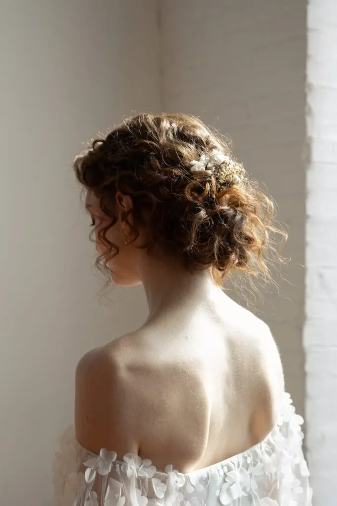 A bride with curly brown hair styled in an elegant updo is seen from the back, showcasing the intricate detail of her hairstyle by Mariage MTL, a vendor in the Wedding Beauty category. The hair is adorned with delicate floral embellishments, complementing the ethereal off-shoulder wedding gown with floral appliqués. The soft lighting and neutral background highlight the romantic and sophisticated bridal look, perfect for a wedding setting.