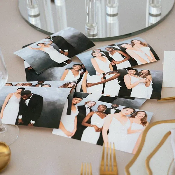 The image shows several printed photographs scattered across a light-colored table, each featuring elegantly dressed wedding guests, including couples and individuals, in formal attire. The backdrop of the photos is a soft, blurred gray, adding a touch of elegance and focus to the subjects. In the foreground, golden cutlery and the edge of fine china are visible, suggesting a refined wedding reception setting. This image represents the services provided by I Party Booth, a photo booth vendor in the wedding category for Mariage Elegant in Canada.