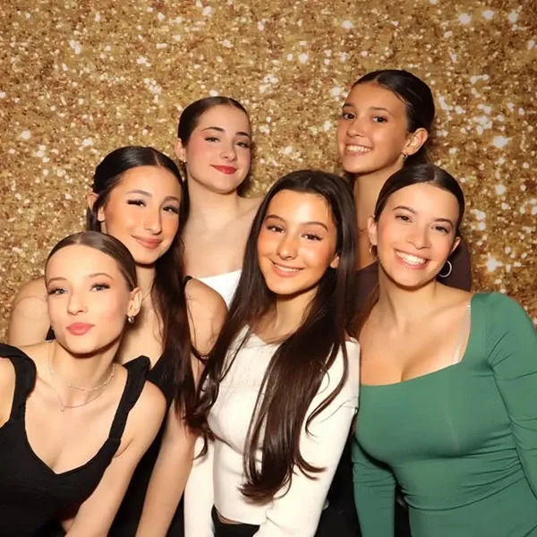 This image features six smiling young women posing together in front of a sparkling gold sequin backdrop, commonly used for creating a glamorous and festive atmosphere at events. They are dressed in assorted elegant outfits, adding to the celebratory feel of the occasion. This setup is part of a photo booth experience offered by I Party Booth, a vendor in the Photo Booth category for wedding planning services provided by Mariage Elegant, Canada. The cheerful and stylish tone makes it perfect for capturing joyful wedding memories.