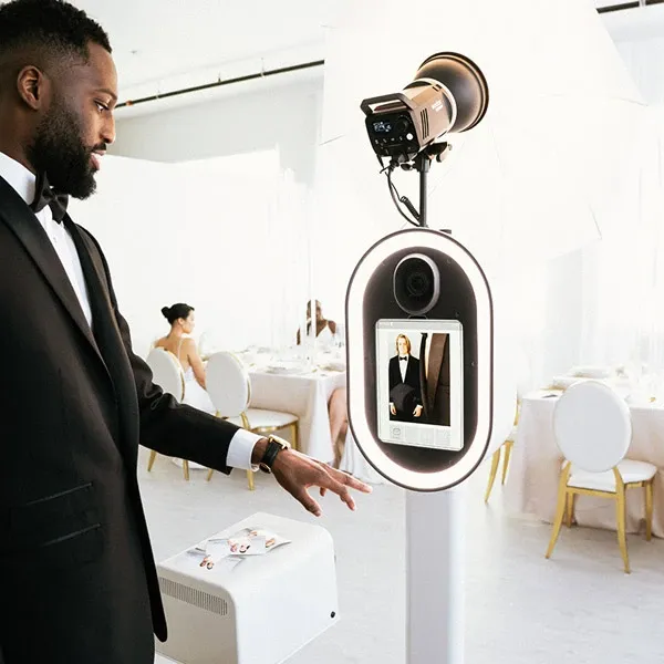 A well-dressed man in a tuxedo interacts with a sleek, modern photo booth machine offered by I Party Booth, part of the Photo Booth category for Mariage Elegant in Canada. The photo booth, elegantly designed with a digital touch screen displaying a preview image, is situated in an elegantly decorated wedding venue with round tables and gold-accented white chairs in the background, highlighting a sophisticated and stylish atmosphere perfect for capturing memorable moments.