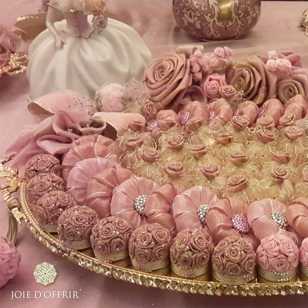 This image features an elegant arrangement of wedding favours by Joie d’Offrir, a vendor specializing in exquisite gifts for weddings. The centerpiece is a lavish golden tray adorned with beautifully crafted pink and gold floral confections, each detailed with intricate designs and sparkling embellishments. The foreground showcases a variety of treats arranged artfully, resembling delicate roses. In the background, a gracefully posed figurine of a bride adds to the romantic and sophisticated ambiance. This display exemplifies the refined and opulent offerings by Joie d’Offrir in the wedding favours category at Mariage Elegant, Canada.